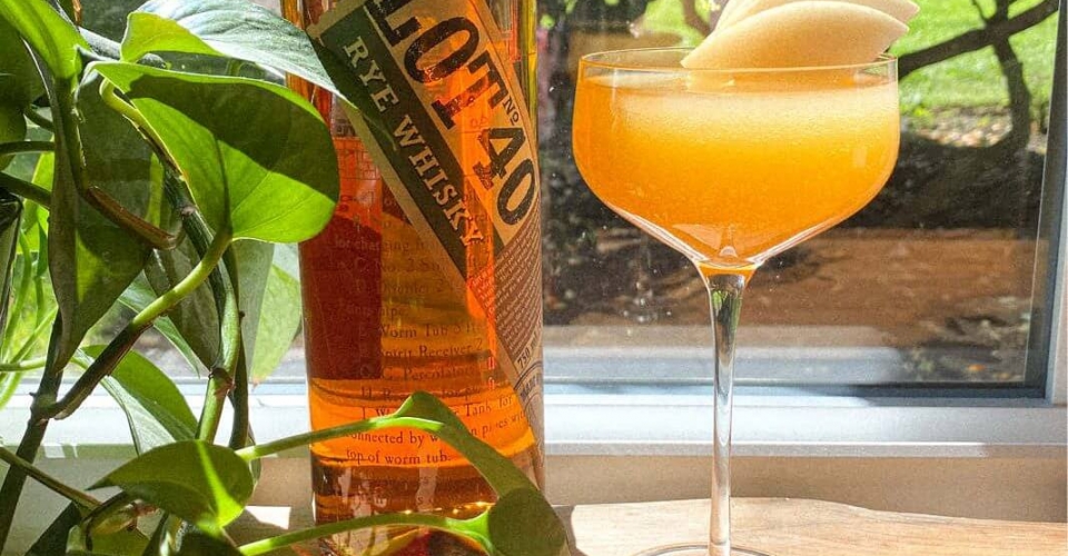 Cocktail with a stone fruit garnish sitting next to a bottle of Lot 40 whiskey.
